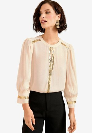 Kurzarm-Bluse in Creme mit goldenen PaillettenAkzenten an Schultern, Ausschnitt und Ärmelbündchen; transparenter Stoff, runder Ausschnitt, lockere Passform.