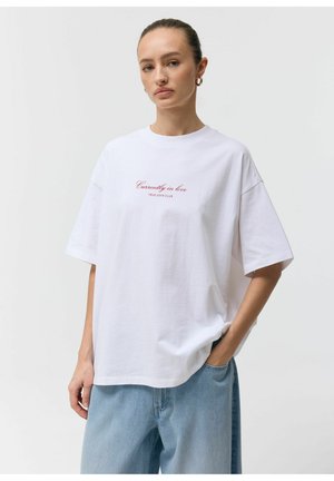 Kobieta w białej, oversize'owej koszulce z napisem „Currently in love TRUE LOVE CLUB”, w jasnoniebieskich dżinsach, stojąca z jedną ręką w kieszeni.