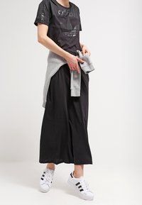 Camiseta deportiva negra con un logotipo brillante, falda maxi negra, suéter gris atado a la cintura y zapatillas blancas con rayas negras.