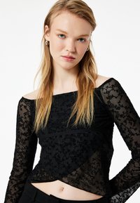Sort langærmet top lavet af gennemsigtig blomsterlacing, med en off-the-shoulder halsudskæring og et cropped design med en struktureret overflade.