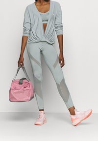 Vaaleanharmaa pitkähihainen paita, siihen sopiva crop-top ja legginssit, joissa on verkkoyksityiskohtia. Vaaleanpunainen urheilulaukku ja kengät vaaleanpunaisilla yksityiskohdilla.