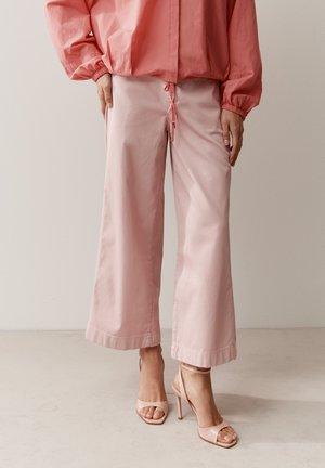 Frau trägt weit geschnittene, hellrosa Hosen, passende offene High Heels und ein korallenfarbenes Langarmoberteil mit elastischen Bündchen.