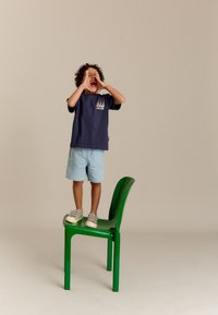 Kind dat op een groene plastic stoel staat, gekleed in een marineblauwe t-shirt met kleurrijke zeilbootafbeeldingen en lichte spijkerbroekshorts, grijze instap schoenen.