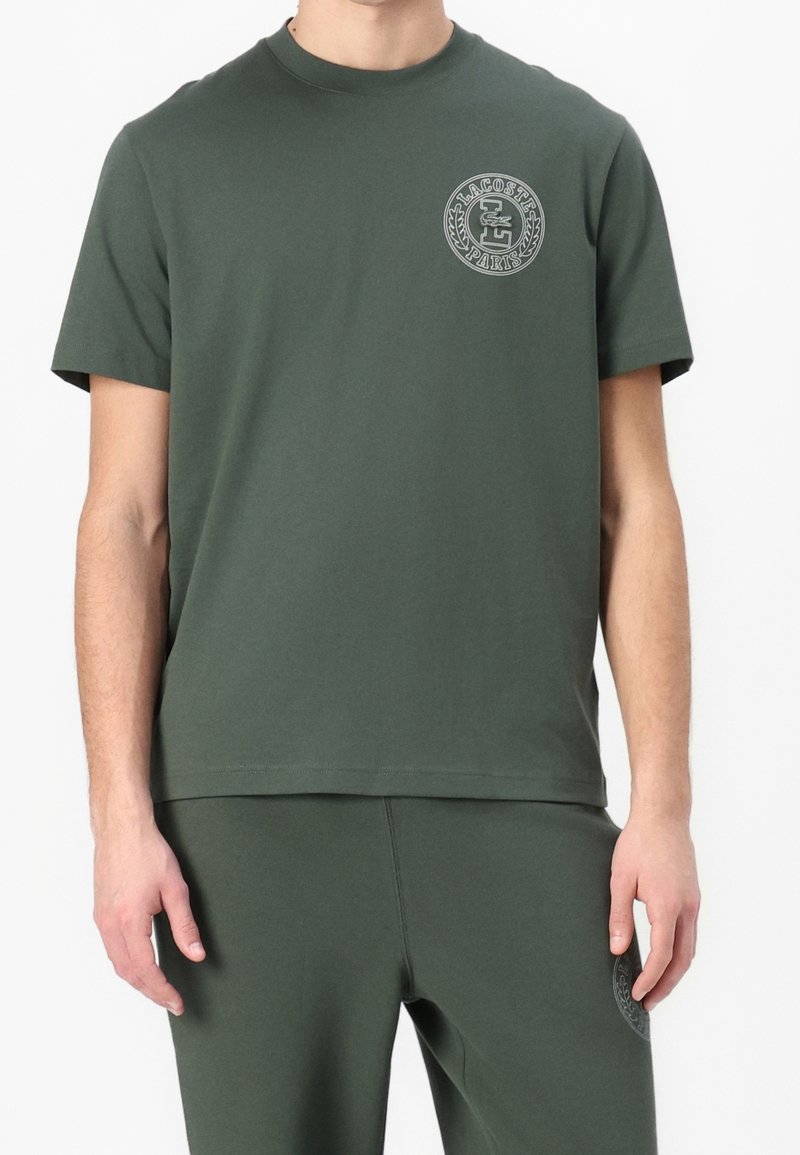 T-shirt en coton vert foncé avec un col rond, des manches courtes, et un logo circulaire gris sur le côté gauche de la poitrine. Texture lisse.