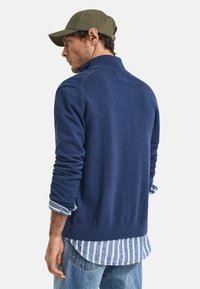Dunkelblauer Strickpullover mit hohem Kragen, ausgestattet mit ribbelten Bündchen und Saum. Trägt ein blau-weiß gestreiftes Hemd darunter. Legere Jeans.