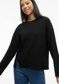 Juodas, ilgomis rankovėmis, apvalia kaklo iškirpte megztinis su laisvu kirpimu ir šoninėmis plyšiais, derinamas su mėlynomis džinsomis. Minkšta tekstūra, minimalistinis dizainas.