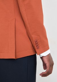 Orange strukturerad kavaj med enkelknäppt design, fyra-knappars manschett och skarpa axelsömmar. Bärs över en vit skjorta.