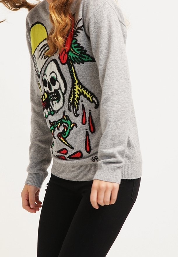 Personne portant un pull gris avec un design de squelette coloré, une griffe d'oiseau et du sang qui coule, associé à un pantalon noir.