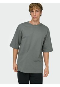 Grijs oversized T-shirt met korte mouwen, ronde hals, gladde textuur en minimale stikdetails. Combinatie met zwarte broek.