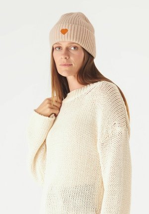 Rosa gerippte Strickmütze mit einem orangefarbenen Herz-Patch. Kombiniert mit einem locker gestrickten cremefarbenen Pullover mit Rundhalsausschnitt und weiten Ärmeln.