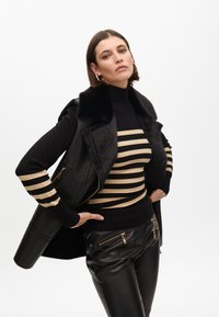 Chaleco de cuero negro con cuello de piel sintética, camiseta de rayas negras y doradas de manga larga, y pantalones de cuero sintético negros con detalles con cremallera.