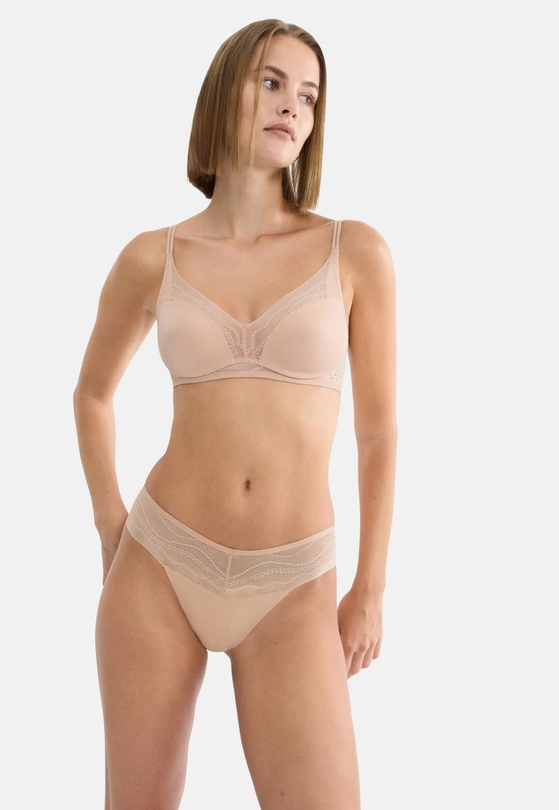Γυναίκα που φοράει σουτιέν σε γυμνό χρώμα και αντίστοιχο εσώρουχο με δαντελένιες λεπτομέρειες, στέκεται μπροστά σε μια απλή ανοιχτόχρωμη επιφάνεια.
