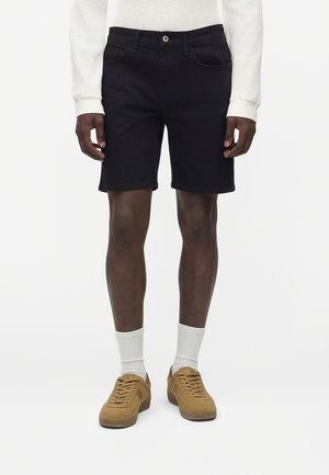 Mand iført sorte denimshorts, hvide ribstrikkede crew-sokker, brune snøresneaksere og en hvid struktureret langærmet sweater, står mod en hvid baggrund.