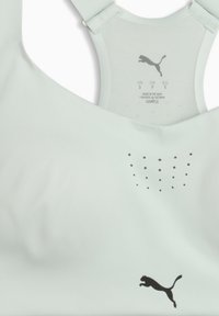 Ανοιχτό γκρι αθλητικό σουτιέν Puma με μαύρο λογότυπο μπροστά και ετικέτα μεγέθους μέσα στη λαιμόκοψη που δείχνει τα μεγέθη US, CA, EU.