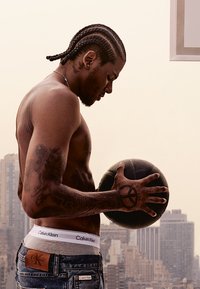 Homme torse nu avec des cheveux tressés tient un ballon de basketball noir. Il porte un jean en denim bleu et une ceinture Calvin Klein, avec des tatouages sur ses bras.