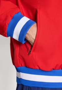 Red jacket with blue ribbed cuffs featuring white stripes. Pocket visible on the left side, textured fabric.