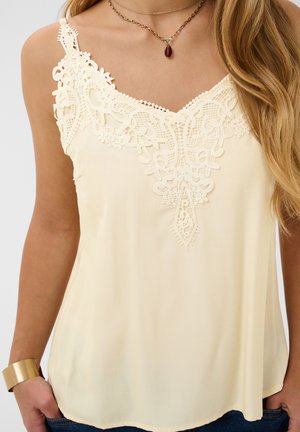 Blusa senza maniche color crema con un profondo scollo in pizzo indossata con collana e braccialetto dorati, mani nelle tasche.