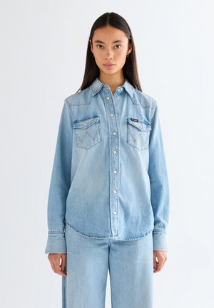 Młoda kobieta z długimi ciemnymi włosami, ubrana w jasnoniebieską koszulę dżinsową na guziki oraz dopasowane dżinsy, stojąca na tle jednolitego białego tła.