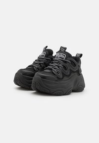 Black sneakers with a chunky profile, textured fabric upper, prominent white laces, and rubber soles featuring a unique tread pattern.
