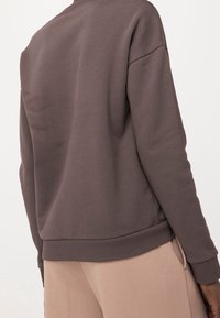 Osoba v hnědé mikině s dlouhým rukávem a béžových kalhotách, zobrazena zezadu na jednoduchém světlém pozadí.