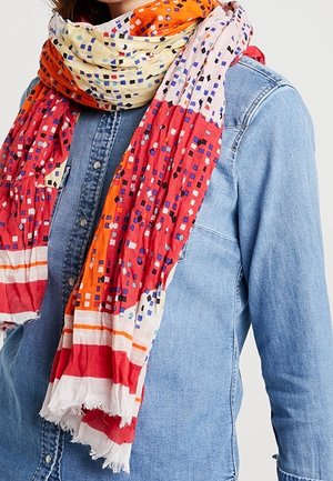 Femme portant une écharpe à motifs rouges, orange et blancs sur une chemise en jean bleu clair boutonnée.