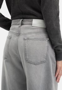 Szare dżinsy z denimu z dwoma tylnymi kieszeniami, skórzaną łatą z logo na talii i luźnym krojem. Tekstura wydaje się miękka i lekko zużyta.