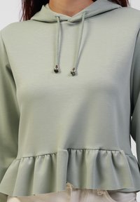 Mintgrønn hettegenser med snor, med volangkant og glatt stofftekstur. Designet inkluderer sølvfargede detaljer.