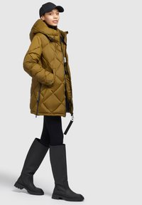 Olive green quilted puffer coat with a hood, side zippers, and a looped pull tab, paired with black knee-high rubber boots.