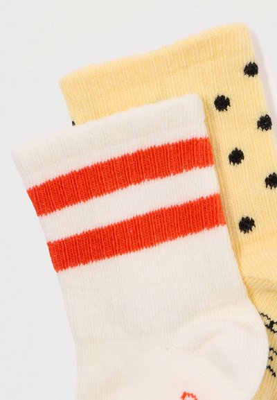Dois pares de meias dobradas, uma branca com duas riscas vermelhas, a outra amarela com bolinhas pretas, contra um fundo branco.