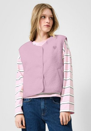 Jeune femme portant un gilet sans manches en polaire violet clair par-dessus une chemise à manches longues rayée rose et blanche, et un jean bleu, regardant vers la droite.