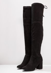 Botas negras por encima de la rodilla hechas de suave ante, con un tacón ancho y puntera redondeada. Presenta una textura lisa y un detalle de tirador en la parte superior.