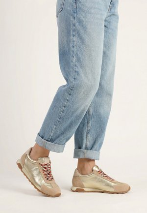Person trägt hellblaue, hochgekrempelte Jeans und metallisch goldene Turnschuhe mit mehrfarbigen Schnürsenkeln, steht vor hellem Hintergrund.