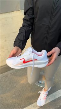 Osoba ubrana w beżowe spodnie i czarną kurtkę trzyma na zewnątrz na chodniku białe i czerwone buty do biegania marki Nike.