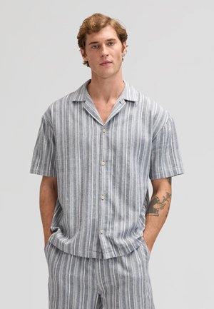 Jonge man met kort krullend haar, gekleed in een grijs-wit gestreept overhemd met korte mouwen en bijpassende broek, handen in de zakken.