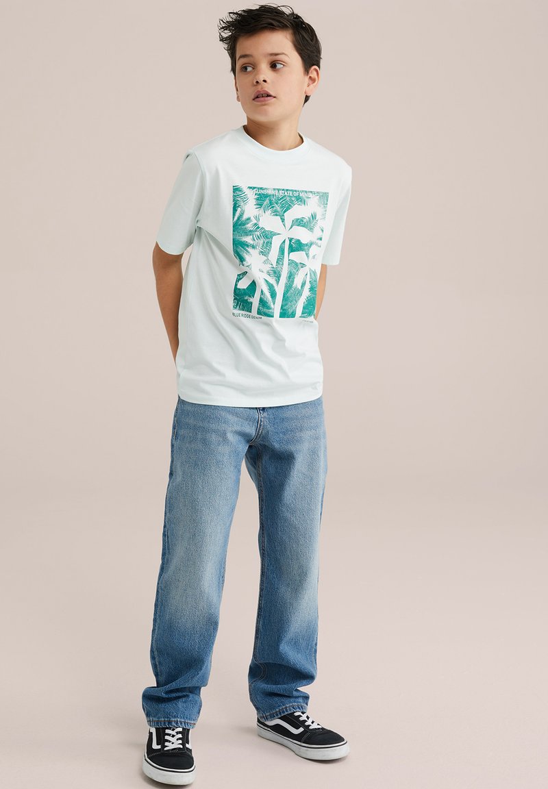 T-shirt grafica azzurro chiaro con motivo di palme, abbinato a jeans in denim chiaro a vestibilità ampia e sneakers nere a bassa cima.