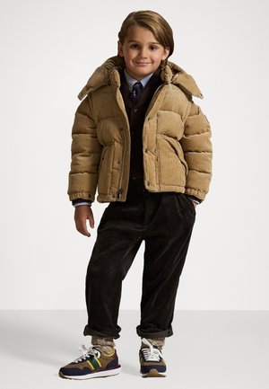 Beige Cordjacke mit großem Kragen, getragen über einem dunklen Weste und einem hellblauen Hemd, kombiniert mit dunklen Cordhosen und gemusterten Turnschuhen.