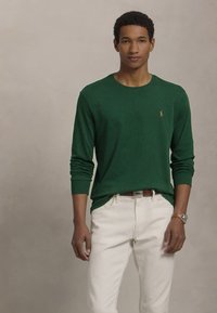 Νεαρός άνδρας φορώντας πράσινο μακρυμάνικο πουκάμισο και λευκά παντελόνια, στέκεται μπροστά σε μια απλή λευκή μπεζ ταπετσαρία.