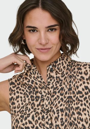 Femme aux cheveux courts et foncés ajustant le col d'une chemise sans manches imprimée léopard, portant des boucles d'oreilles créoles argentées, sur un fond uni.