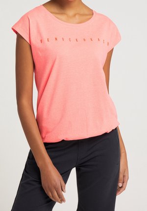 Jeune personne portant un t-shirt corail rose à manches courtes avec le texte "Venice Beach" et un pantalon noir, debout devant un fond uni.