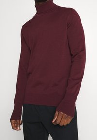 Pull à col roulé bordeaux en tricot léger. Doté de poignets et d'ourlet côtelés, d'une coupe ajustée et de transitions d'épaule sans couture.