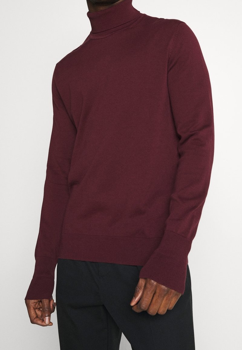 Pull à col roulé bordeaux en tricot léger. Doté de poignets et d'ourlet côtelés, d'une coupe ajustée et de transitions d'épaule sans couture.