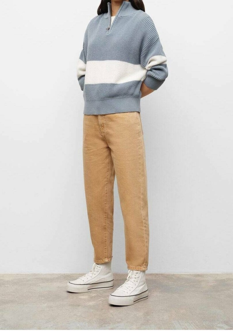 Personne portant un pull rayé bleu et blanc, un jean taille haute beige et des baskets blanches montantes, debout contre un mur uni.