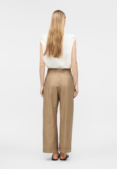 Γυναίκα με μακριά ξανθά μαλλιά στέκεται με την πλάτη γυρισμένη, φορώντας λευκό αμάνικο τοπ, μπεζ φαρδιά παντελόνια με ψηλή μέση και μαύρα σανδάλια.