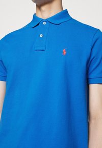 Niebieska koszulka polo z teksturowanej bawełny, z klasycznym kołnierzykiem, dwoma guzikami z przodu i małym czerwonym haftowanym logo na piersi.
