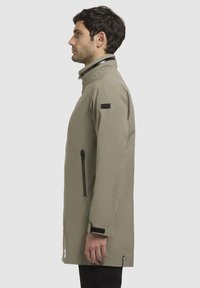 Khaki vandtæt jakke med høj krave, sort lynlås, sidelommer og velcro-manchetter. Slank silhuet.