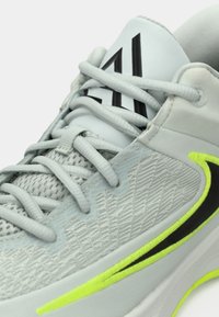 Lys grå sportsko med mesh-overdel, grå snørebånd, sort "AI" logo på tungen og neon gul og sort Nike swoosh på siden.