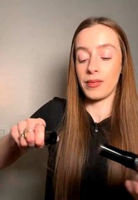 Femme aux longs cheveux raides, les coiffant avec un fer à lisser noir sur un fond beige uni.