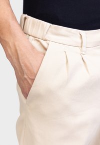 Hand in zak van beige hooggetailleerde broek met plooien, gedragen met een aansluitend zwart topje.