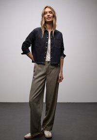 Femme portant une veste en jean foncé, une chemise blanche, un pantalon large gris et des baskets blanches, debout devant un fond clair uni.