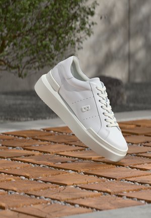 Scarpa da ginnastica bianca bassa con lacci e dettaglio "B" in rilievo, sospesa sopra piastrelle di pietra marrone all'aperto vicino a fogliame verde.